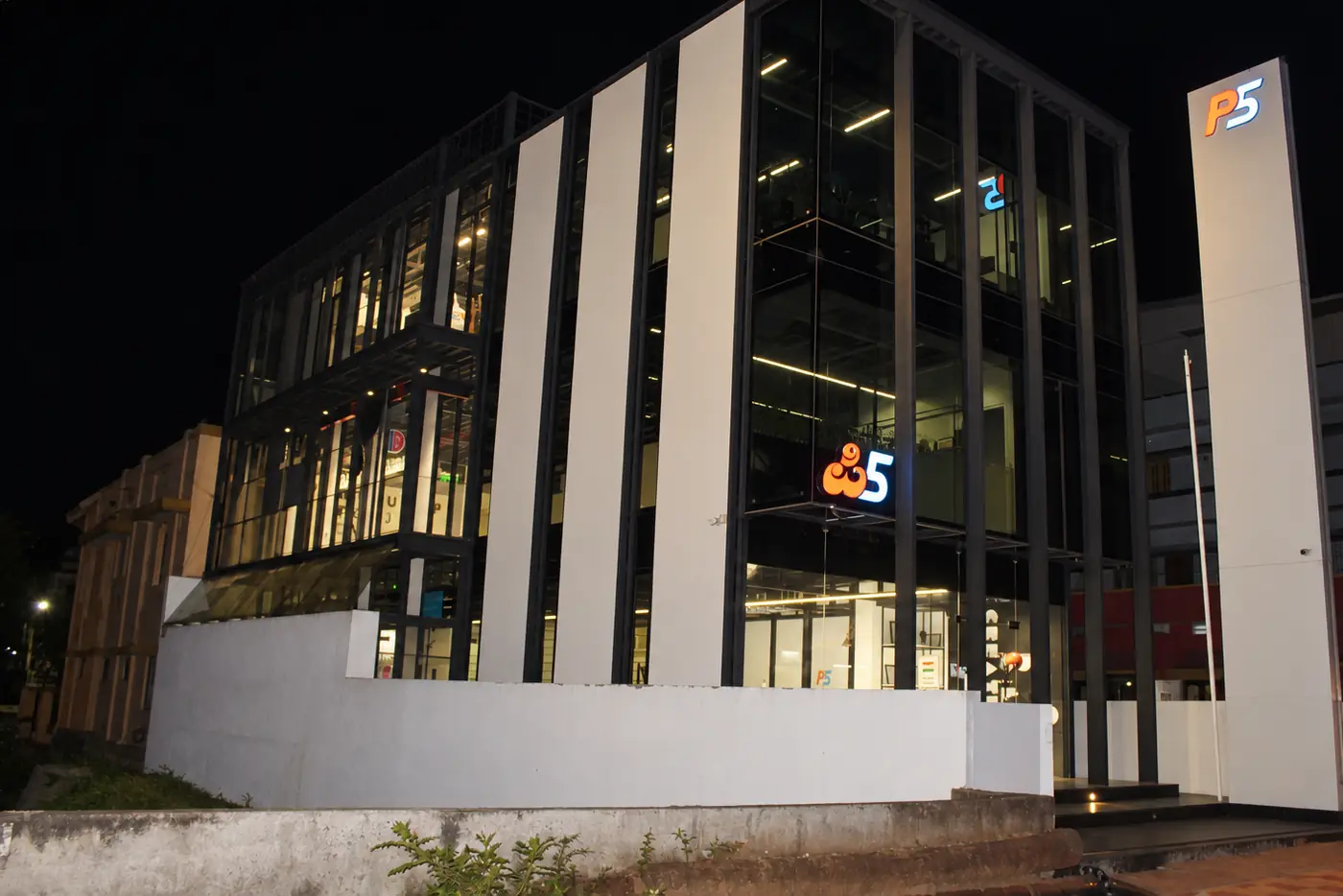 The P5 India building at night: the glass front lit from inside, the P5 logos glowing on the front and on the pylon