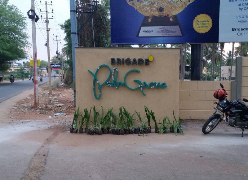 Brigade Palm Grove name in green letters on a stone-clad compound wall beside the road
