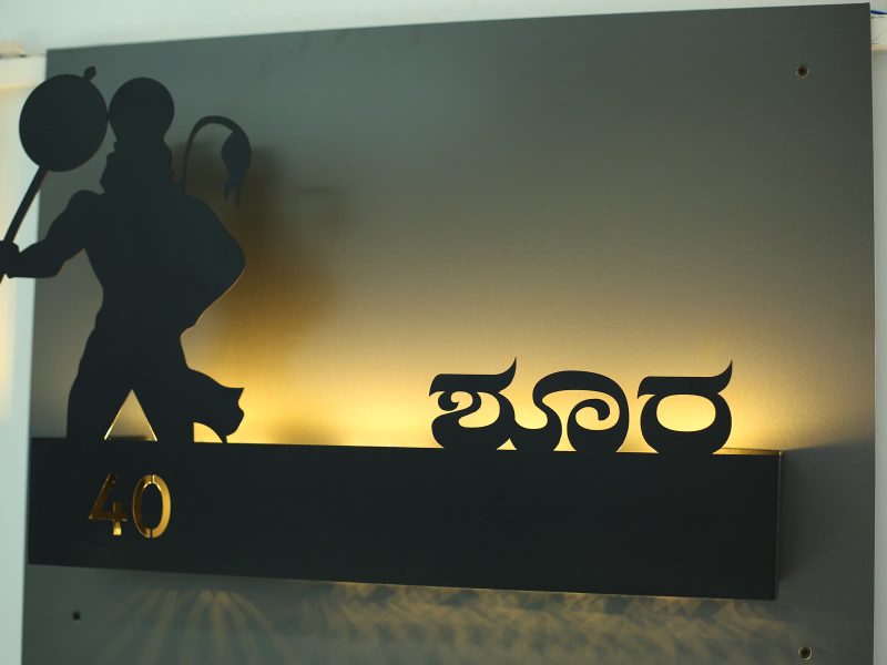Black name plate with Kannada letters and a cut-out figure, lit from behind so the wall glows